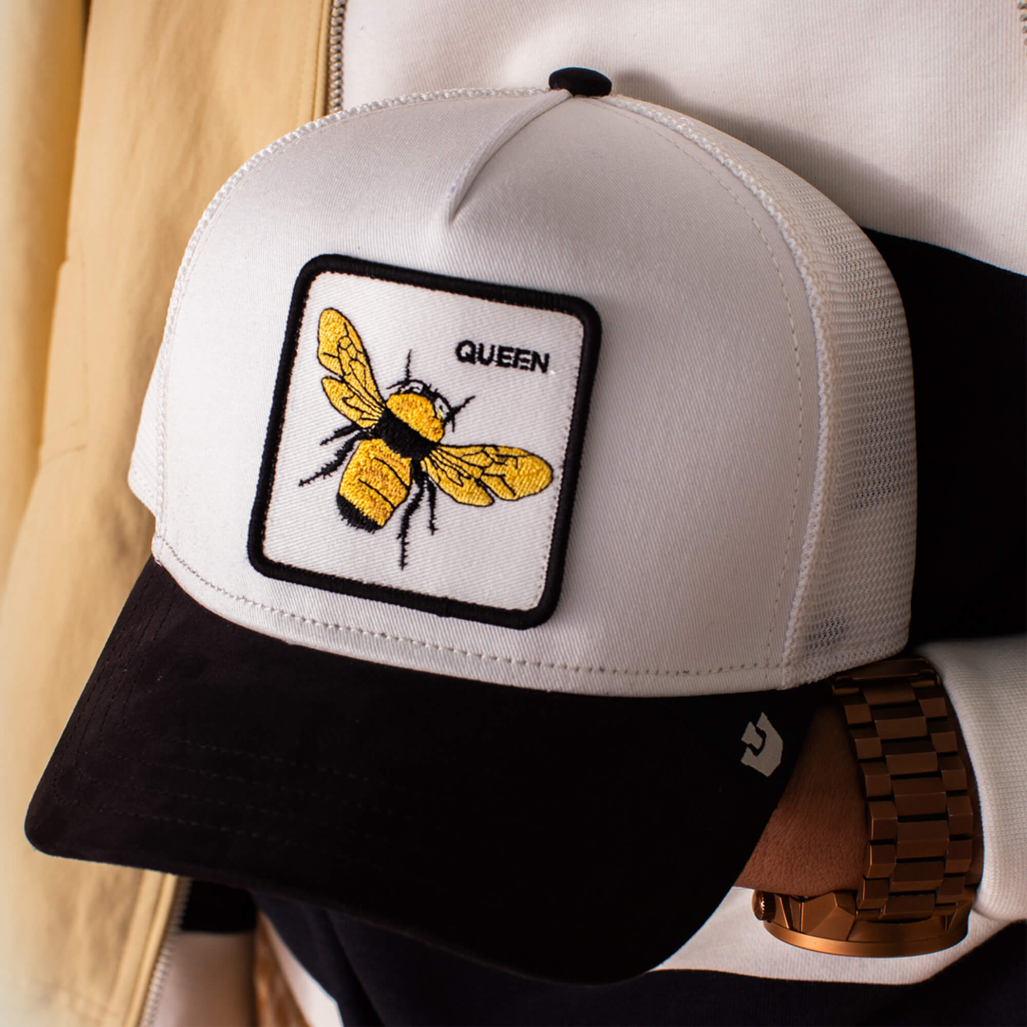 The Goorin Bros. Suede Bee is a white and black trucker hat with a black microsuede brim, featuring a Queen patch adorned with an embroidered yellow bee.
