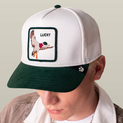 A person wears the Goorin Bros. Suede Duck trucker hat with a white crown, green suede brim, and an embroidered patch featuring a flying duck and "LUCKY" on the front.