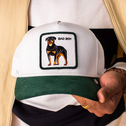 A person holds the Goorin Bros. Suede Bad Boy trucker hat, which is white and green and features a bold Rottweiler patch with "BAD BOY" above the dog.