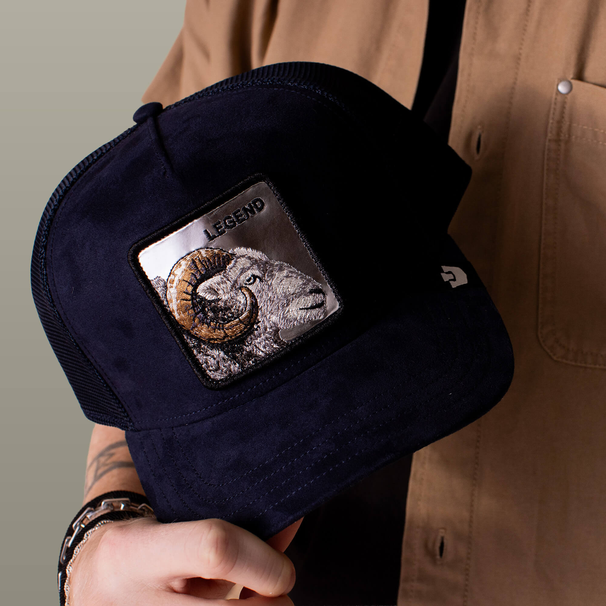 A person holds a navy Goorin Bros. Silver Legend trucker hat with an embroidered ram patch labeled "LEGEND," while wearing a tan shirt and bracelets.