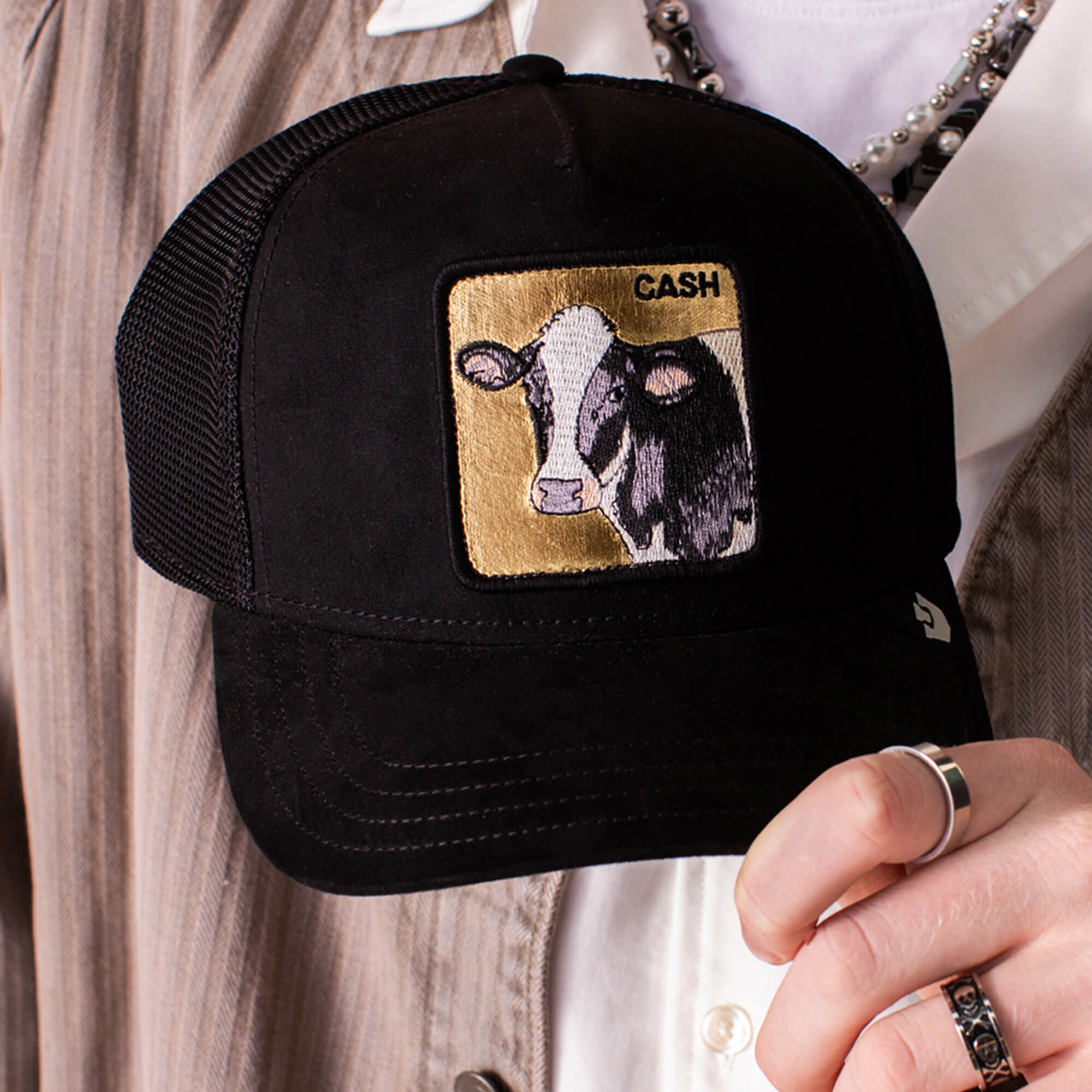 A person holds a black Goorin Bros. Golden Cow trucker hat with a black suede brim and a front patch displaying an illustrated cow and the word CASH.