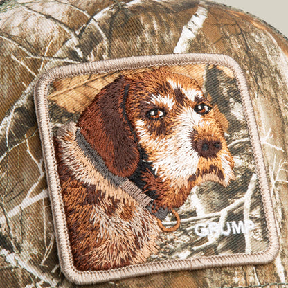 The Realtree Edge Grump Dog by Goorin Bros. features camo fabric with a sewn patch showing an embroidered brown and white dog’s face and "GRUMP" in bold white letters.