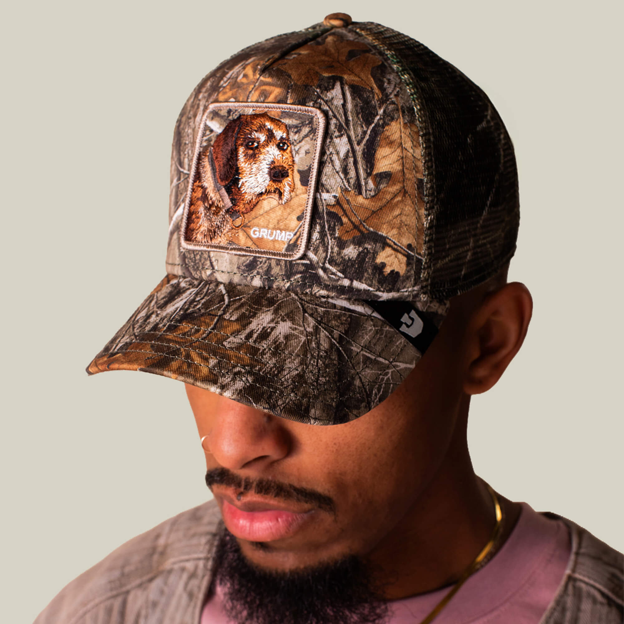 A man in a Goorin Bros. Realtree Edge Grump Dog cap, featuring an embroidered dog patch, looks down with grumpy energy against a plain background.