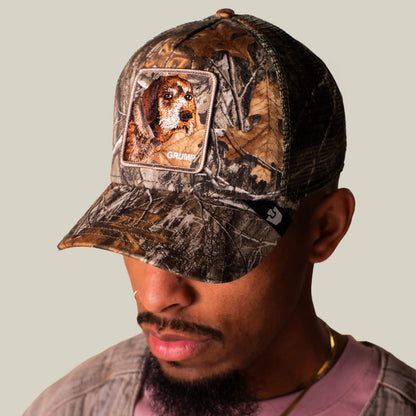 A man in a Goorin Bros. Realtree Edge Grump Dog cap, featuring an embroidered dog patch, looks down with grumpy energy against a plain background.