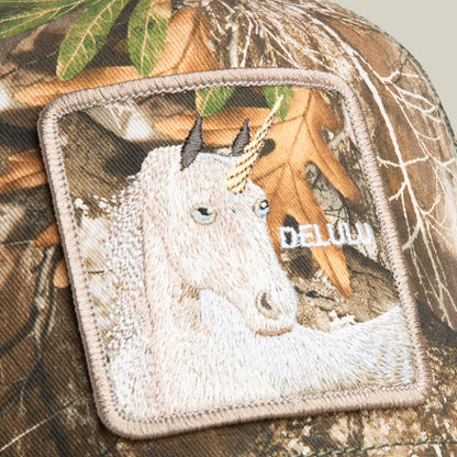 Close-up of the Goorin Bros. Realtree Edge Delulu Unicorn hat, showing camo fabric with an embroidered unicorn patch—white unicorn, golden horn, and the word DEJAVU partially visible.