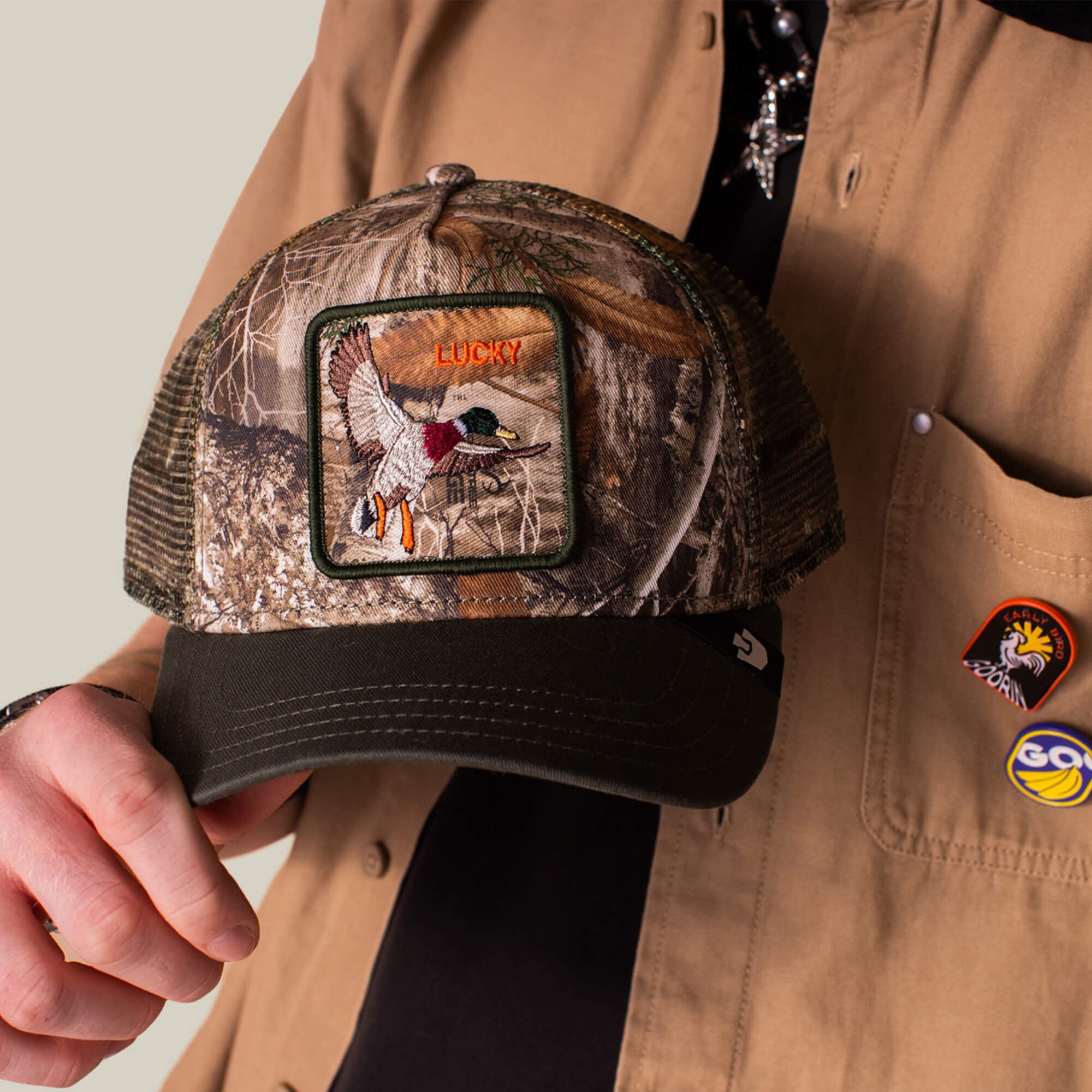 A person in a tan shirt with pins holds the Goorin Bros. Realtree Edge Lucky Duck cap, which features an embroidered duck patch and "LUCKY" on the front.