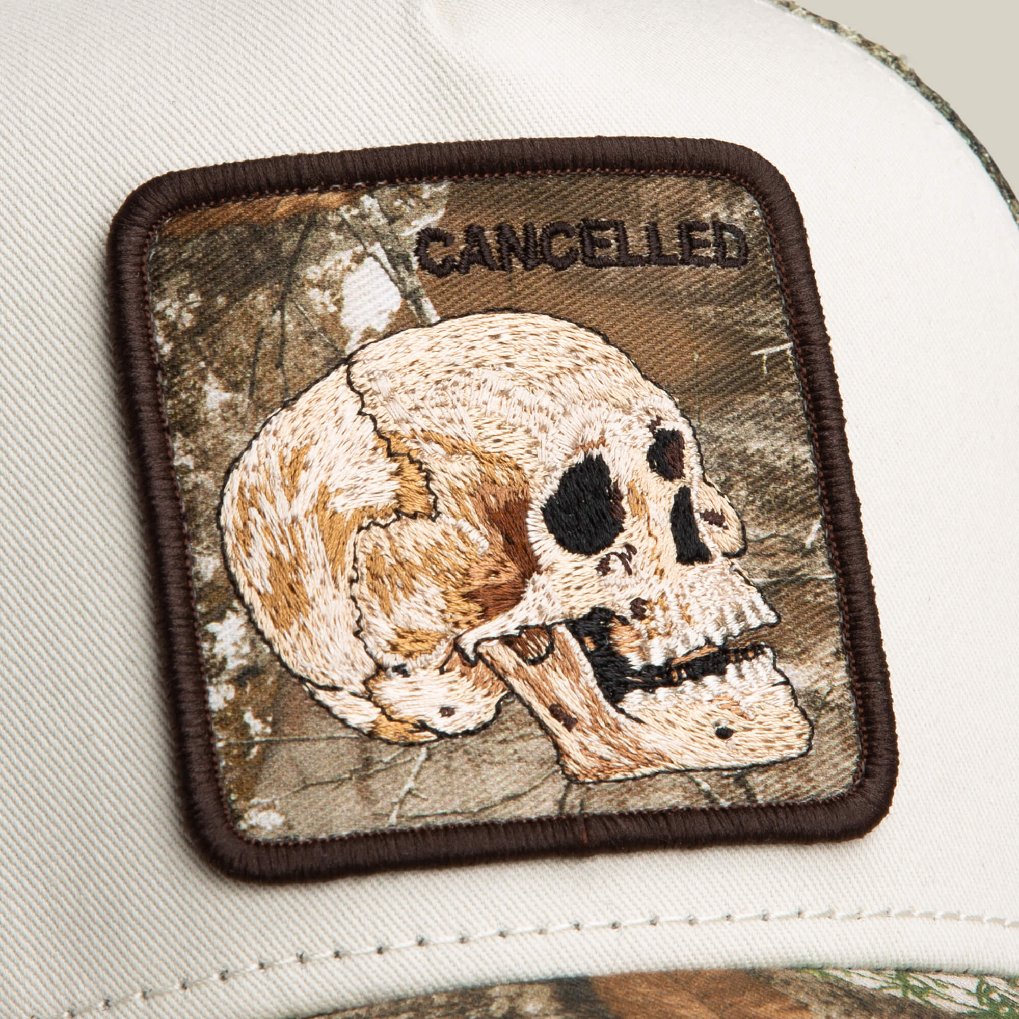A close-up of the Goorin Bros. Realtree Edge Cancelled Skull patch on a cap, featuring a skull and "CANCELLED" text against a Realtree Edge camo background.