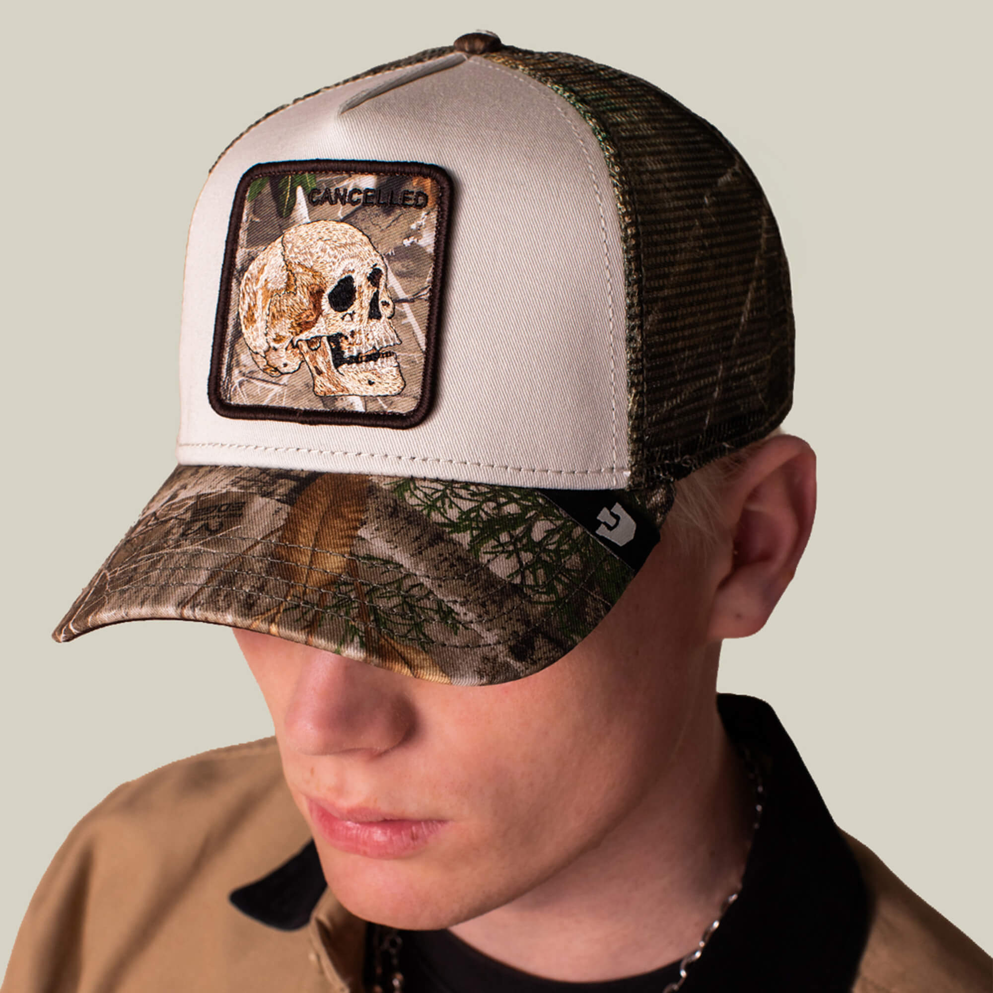 A person wearing the Goorin Bros. Realtree Edge Cancelled Skull trucker hat, featuring a white and camo colorway with a skull patch and "CANCELLED" text, looks downward against a plain background.