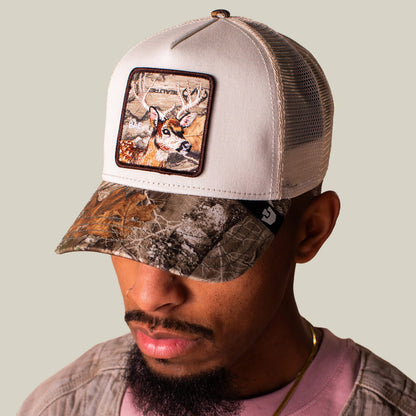 A man wears the Goorin Bros. Realtree Edge Bae Doe hat—a beige mesh cap with a Realtree Edge camo visor and a deer patch—while looking down, sporting a nose ring.