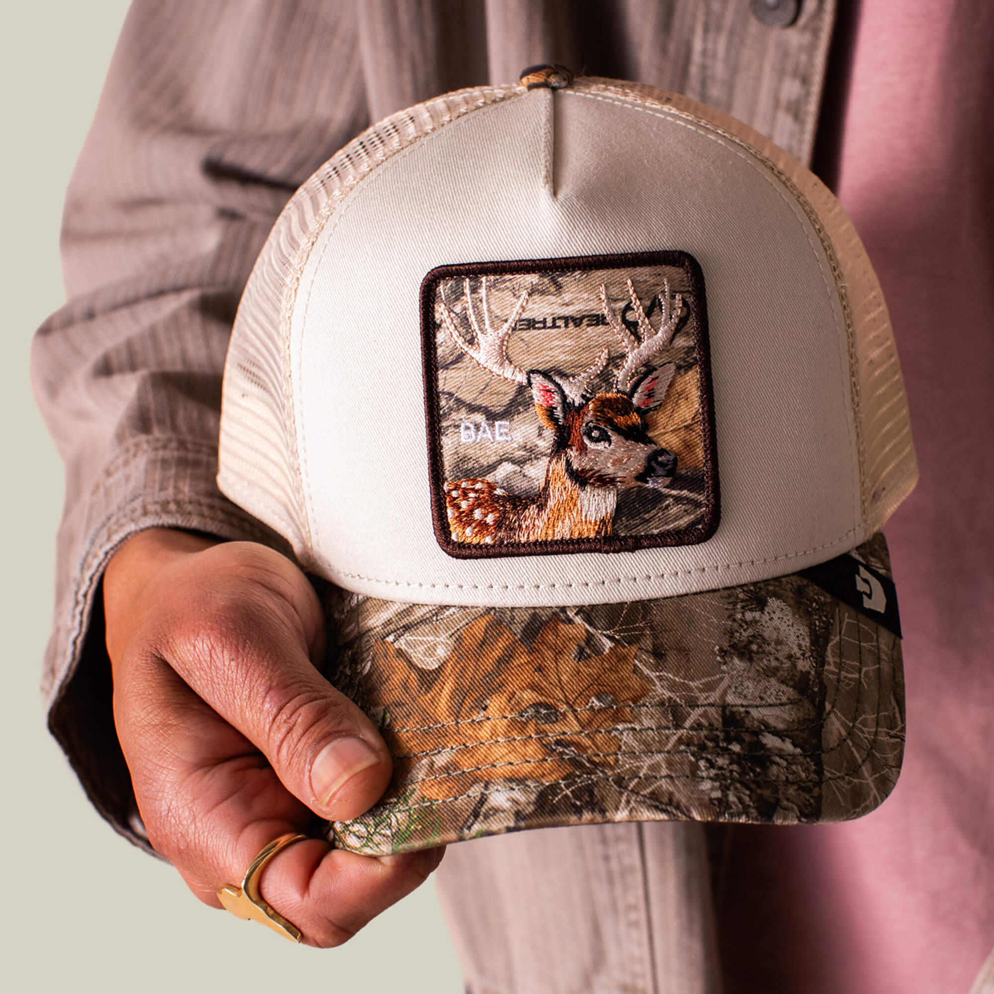 A person holds the Goorin Bros. Realtree Edge Bae Doe hat, featuring a camo brim and a front deer patch with striking antlers.