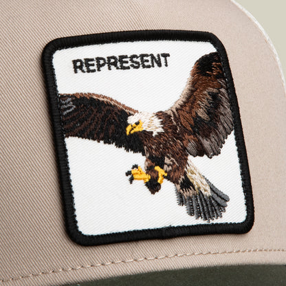A close-up of the Goorin Bros. Represent Eagle Trucker hat, showcasing a patch with an embroidered eagle in flight and "REPRESENT" above it.