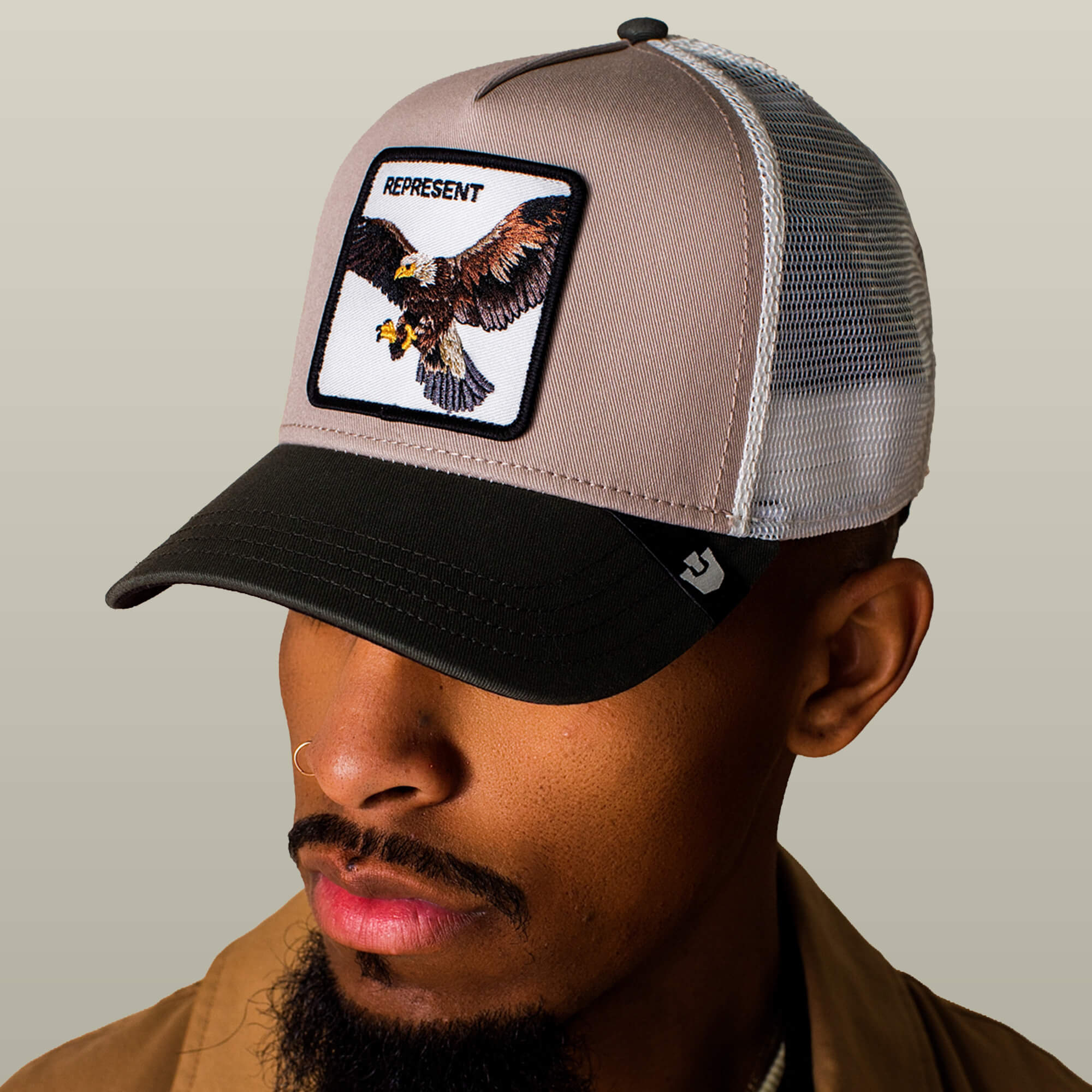 A man wears the Goorin Bros. Represent Eagle Trucker hat, featuring an eagle patch and "REPRESENT" on the front, set against a plain background.