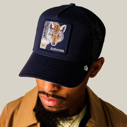 A man in a Goorin Bros. Mirage Survivor navy trucker hat with a fox patch, tan jacket, and white shirt looks downward against a plain background.