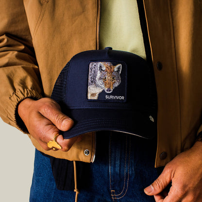 A person in a brown jacket and blue jeans holds the Goorin Bros. Mirage Survivor cap, which features an embroidered fox patch and the word SURVIVOR.