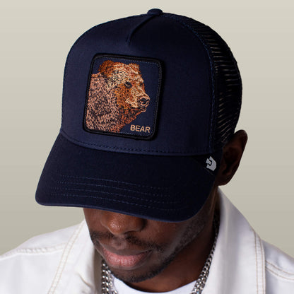 A man wearing the Goorin Bros. Bear Trucker cap, featuring a bear patch and "BEAR" embroidered on the front, looks downward.