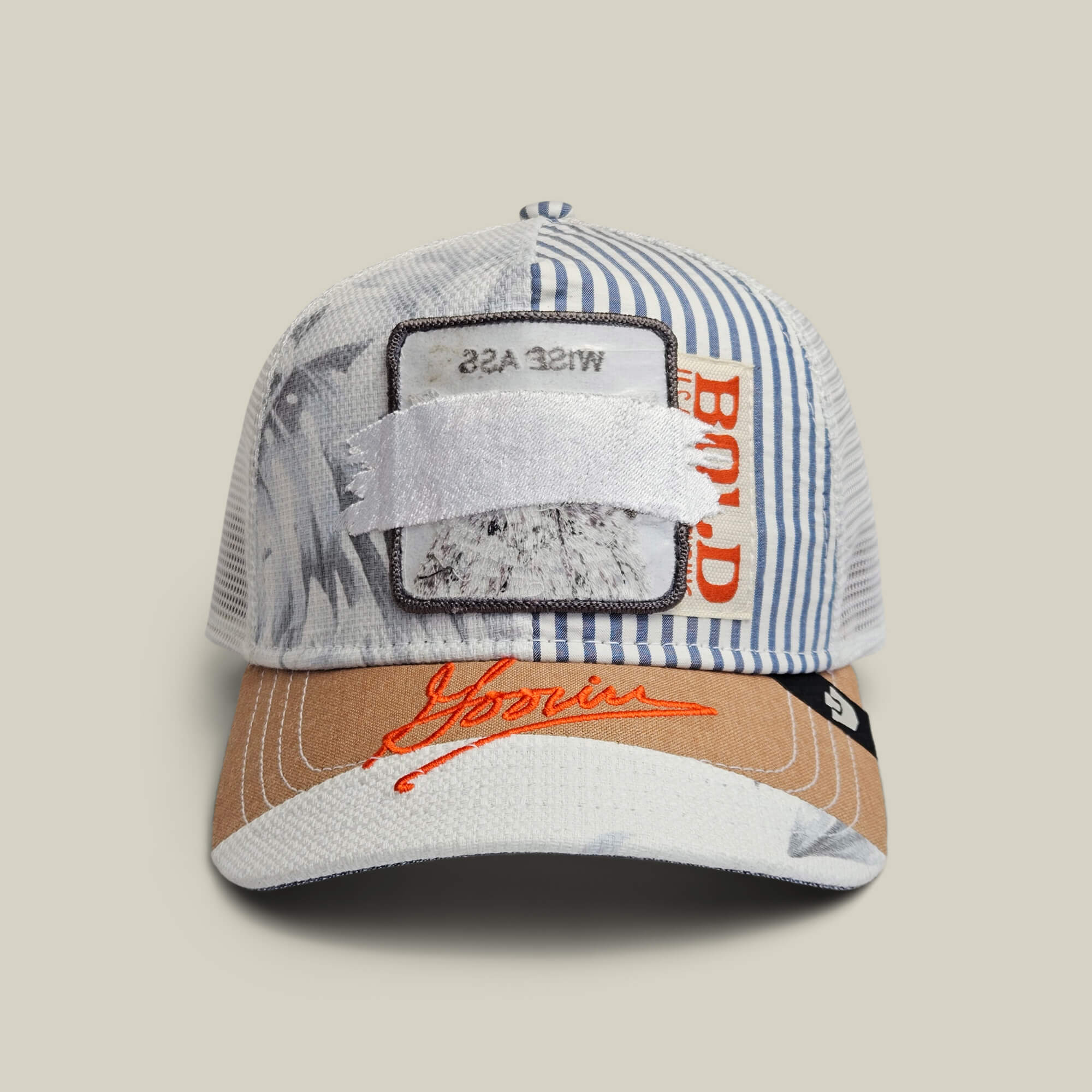 The Goorin Bros. Goorin Rework Trucker in AIR is a limited edition multicolored cap featuring blue stripes, orange text, a brown brim, and a white patch with WISE ASS partially covered plus red embroidered branding on the front.