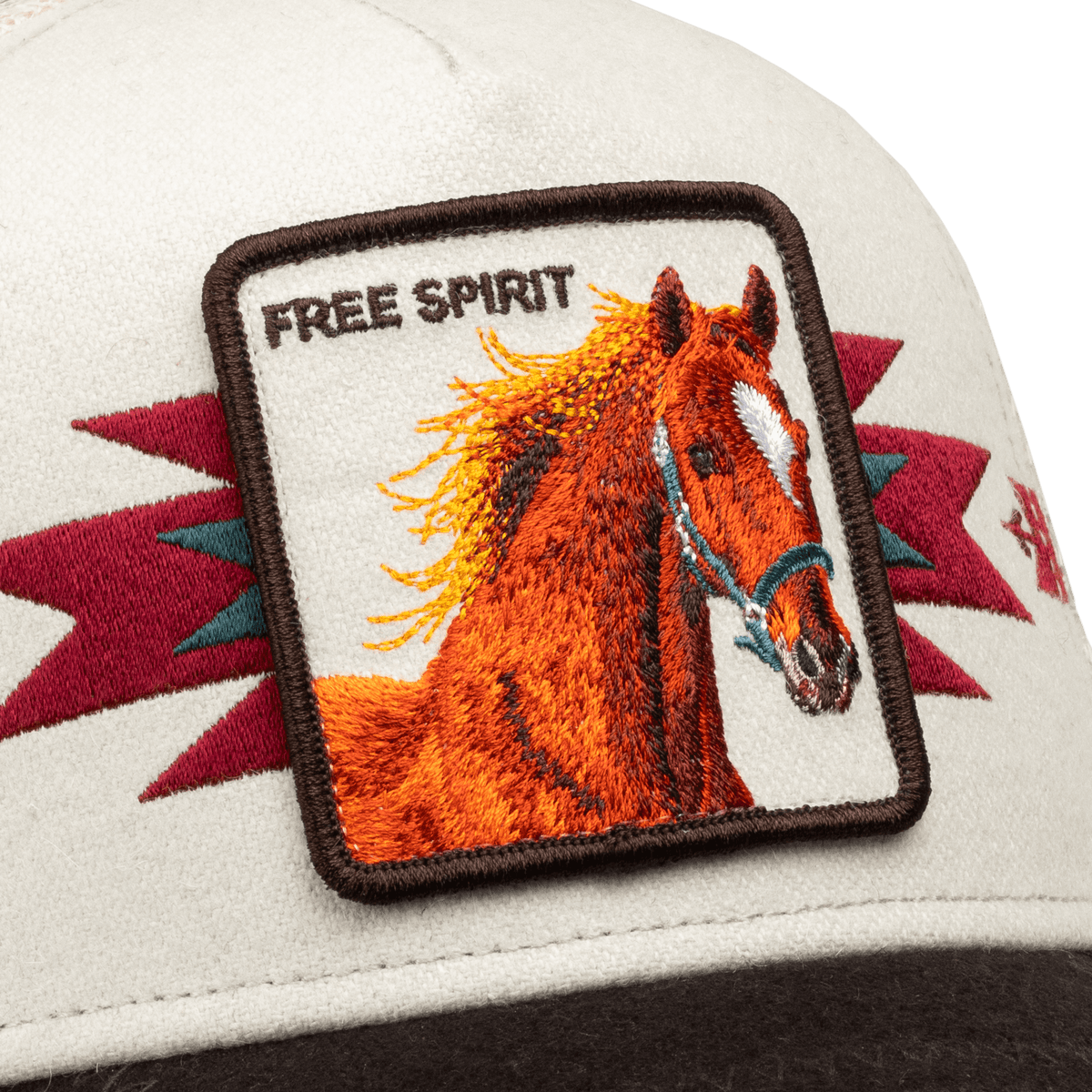 The Canyon Free Spirit trucker hat by Goorin Bros features a cream-colored front with an embroidered orange horse patch and burgundy geometric accents, shown in close-up detail against a black background.