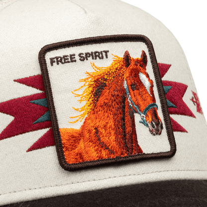 The Canyon Free Spirit trucker hat by Goorin Bros features a cream-colored front with an embroidered orange horse patch and burgundy geometric accents, shown in close-up detail against a black background.
