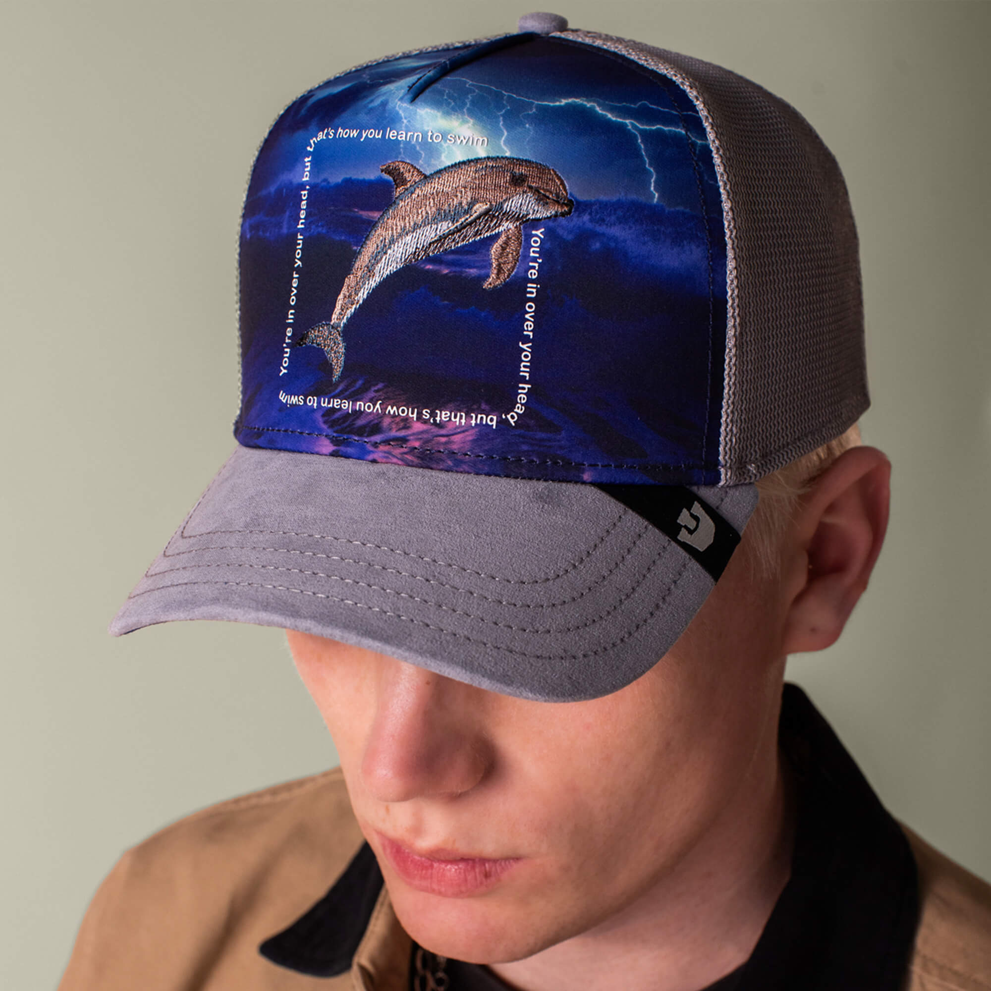 A person wearing the Goorin Bros. "Through the Storm - Dolphin" cap, which features a dolphin leaping from water amid lightning, with text curved around the dolphin.