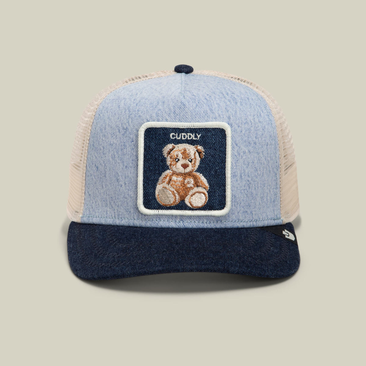 The Goorin Bros. Denim Cuddly trucker hat features a light blue denim front, cream mesh back, navy brim, and embroidered teddy bear patch on a dark navy label, displayed against a neutral studio background.