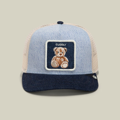 The Goorin Bros. Denim Cuddly trucker hat features a light blue denim front, cream mesh back, navy brim, and embroidered teddy bear patch on a dark navy label, displayed against a neutral studio background.