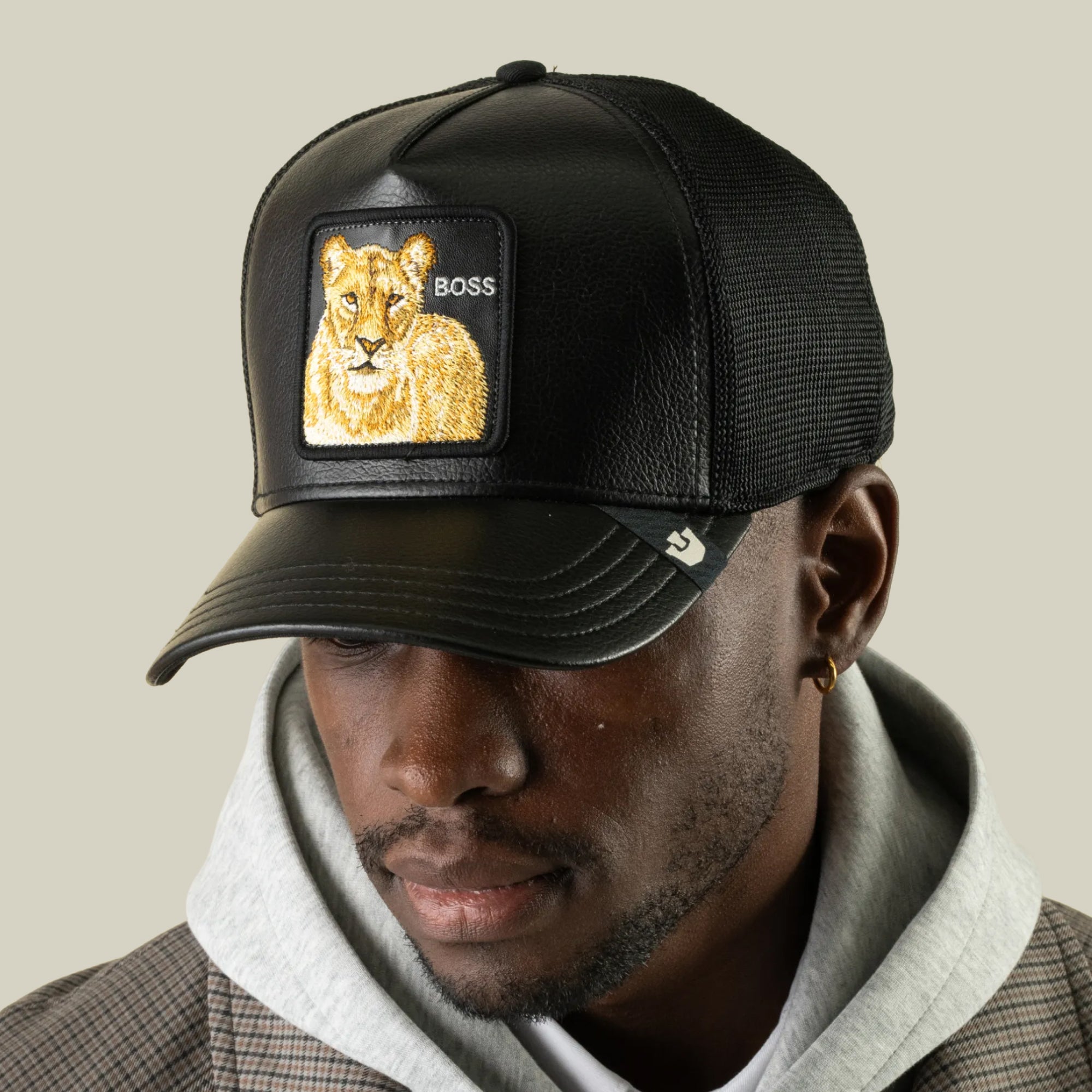 A man wearing the Goorin Bros. “Faux Leather Lioness” black faux-leather trucker hat with a breathable mesh back and a lion patch reading “BOSS,” layered over a gray hoodie and plaid blazer, looks down.