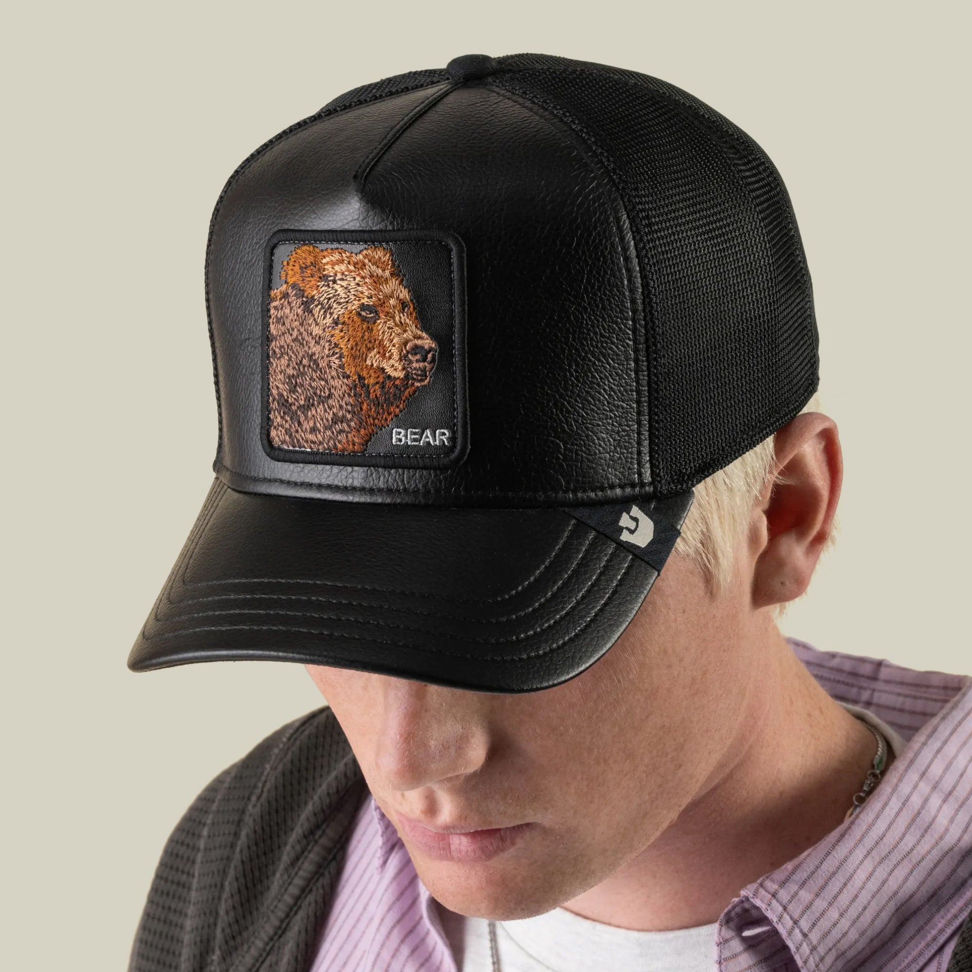 A person wearing the Goorin Bros. Faux Leather Bear trucker hat, featuring a black faux leather front with an embroidered grizzly patch, breathable mesh sides, a leather brim, and a small logo tag on the side, looking down.