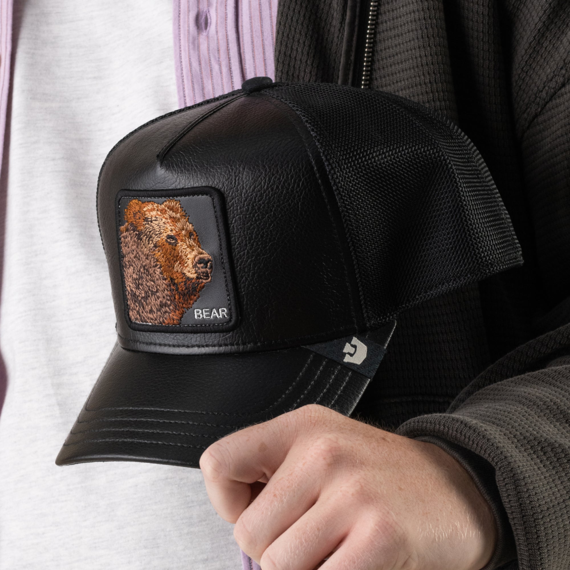A person holds the “Faux Leather Bear” trucker baseball cap by Goorin Bros., featuring black faux leather, breathable mesh sides, and an embroidered grizzly bear patch with the word “BEAR.”.