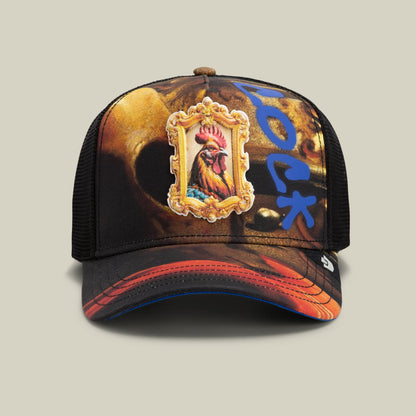 The Penitent Magdalene by Goorin Bros is a black mesh trucker hat featuring an ornate gold-framed embroidered rooster patch at the center, with blue symbols and red gradient accents along the brim, shown on a neutral background.