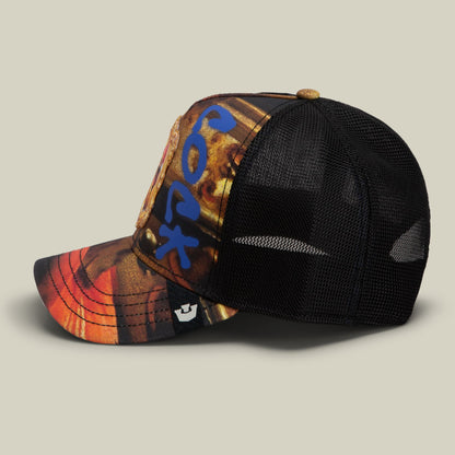 The Penitent Magdalene by Goorin Bros. is a trucker hat featuring a black mesh back and a colorful front panel with blue, orange, gold, and brown tones, displayed against a neutral background.