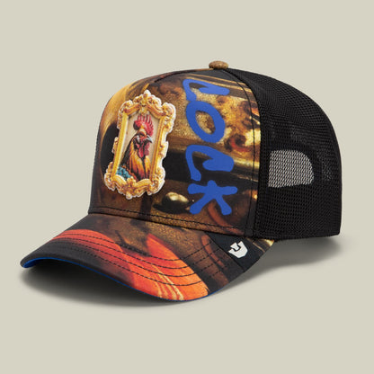 The Penitent Magdalene by Goorin Bros. is a trucker hat with a black mesh back and a printed front panel in brown, orange, and gold tones featuring a rooster emblem in an ornate frame and blue graphic elements, with a red and orange ombre bill.