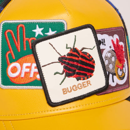 The Goorin Bros. Bugger trucker hat features a bright yellow leather front panel with embroidered animal patches including a red beetle, displayed against a colorful green and purple background in a studio product shot.