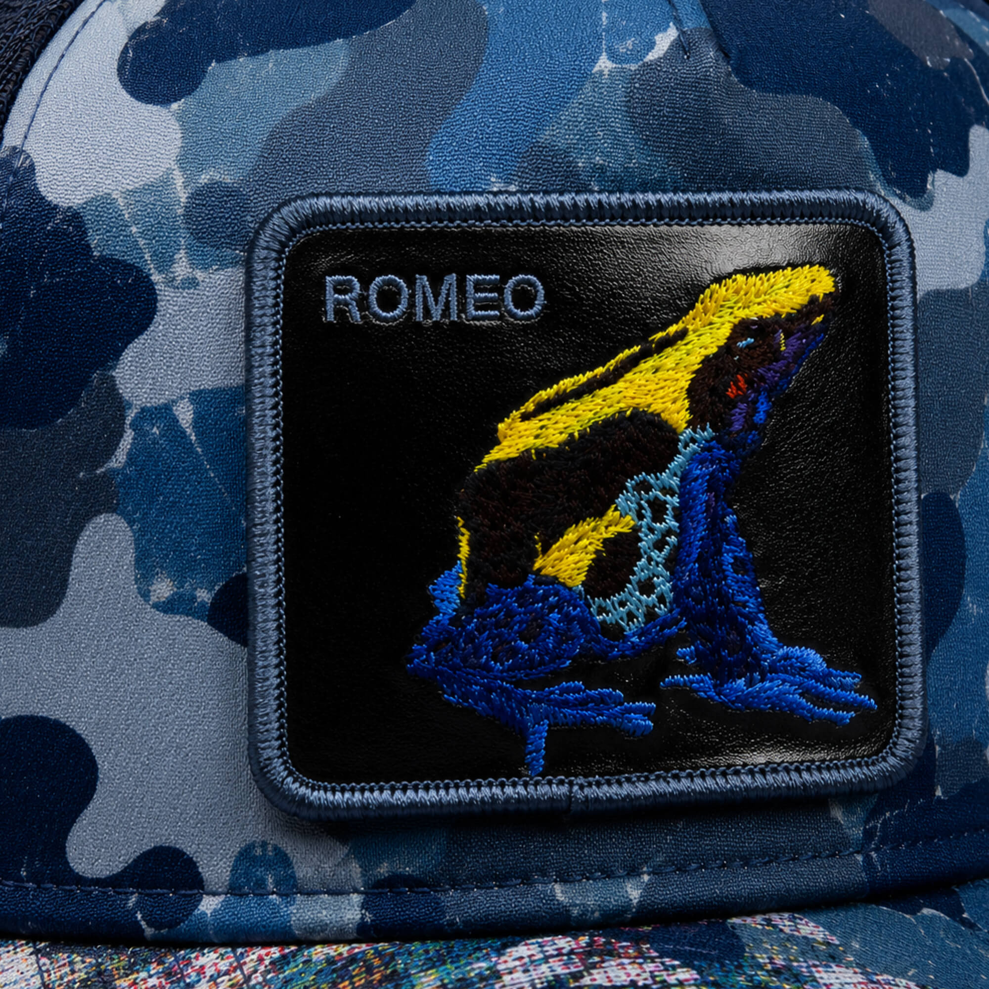 The Warfare Art Thou camo trucker hat by Goorin Bros. showcases a ROMEO poison dart frog patch and an embroidered yellow and blue frog, combining bold style with rugged appeal.