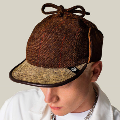 A person wears the Goorin Bros. Frazier Lake brown wool trapper cap with ear flaps and a bow, paired with a white shirt and silver chain necklace.