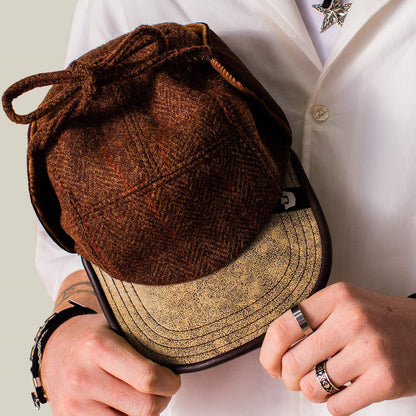A person in a white shirt holds the Goorin Bros. Frazier Lake brown wool trapper cap with ear flaps and a beige brim, displaying rings and bracelets on their hands.