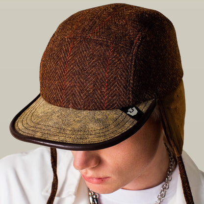 A man looks down wearing the Goorin Bros. Frazier Lake brown wool trapper winter cap with ear flaps and a textured beige brim, paired with a white shirt and chain necklace.