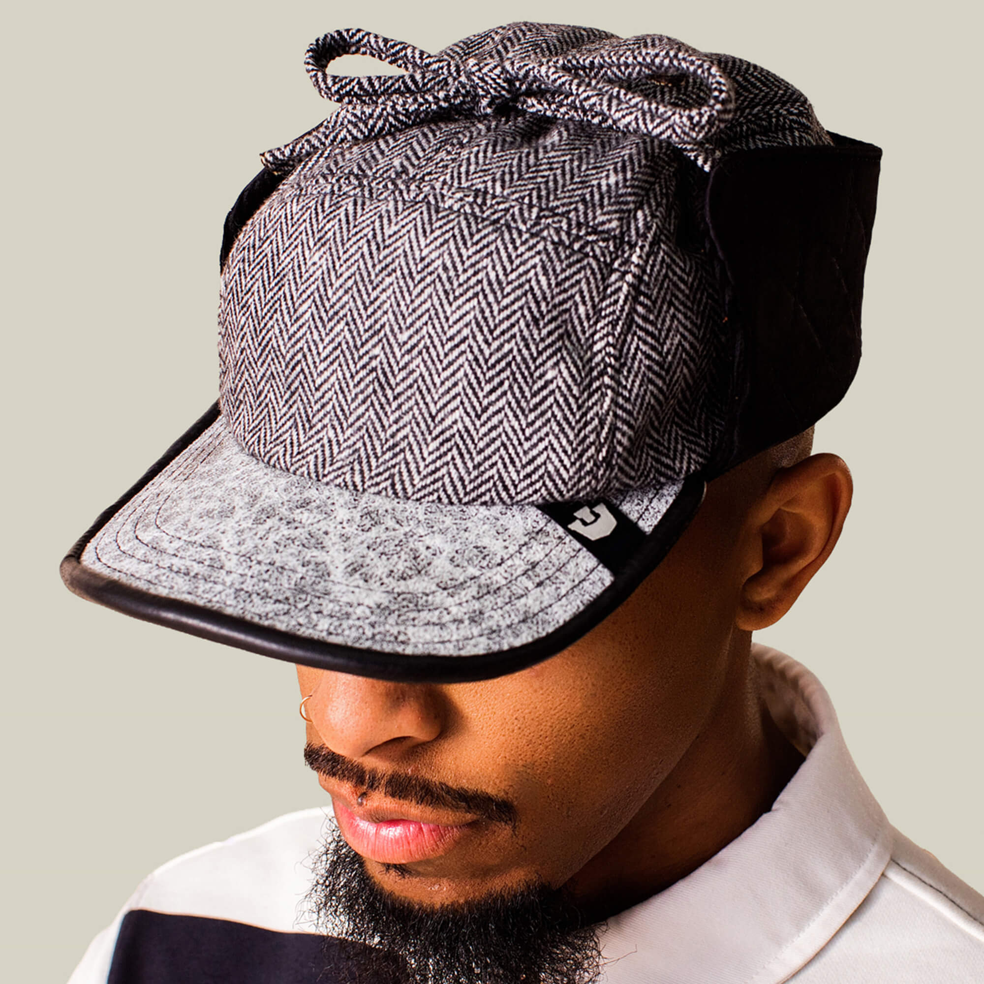 A person wearing the Goorin Bros. Frazier Lake wool trapper cap with a herringbone pattern, black side panels, ear flaps, and a bow on top looks down while dressed in a collared shirt.