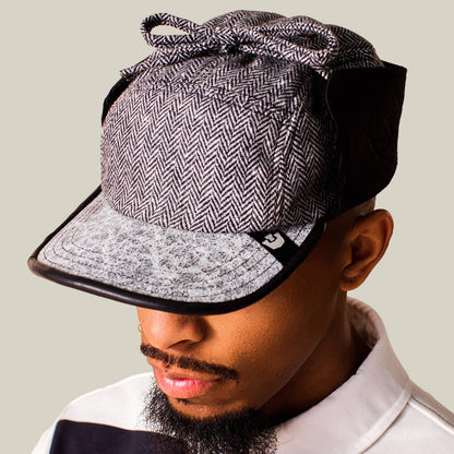 A person wearing the Goorin Bros. Frazier Lake wool trapper cap with a herringbone pattern, black side panels, ear flaps, and a bow on top looks down while dressed in a collared shirt.