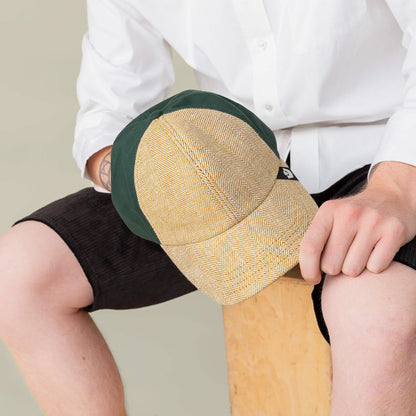 A person in a white shirt and dark shorts sits on a wooden stool, holding the Goorin Bros. Zig Zag Tailored Baseball cap in tan and green.