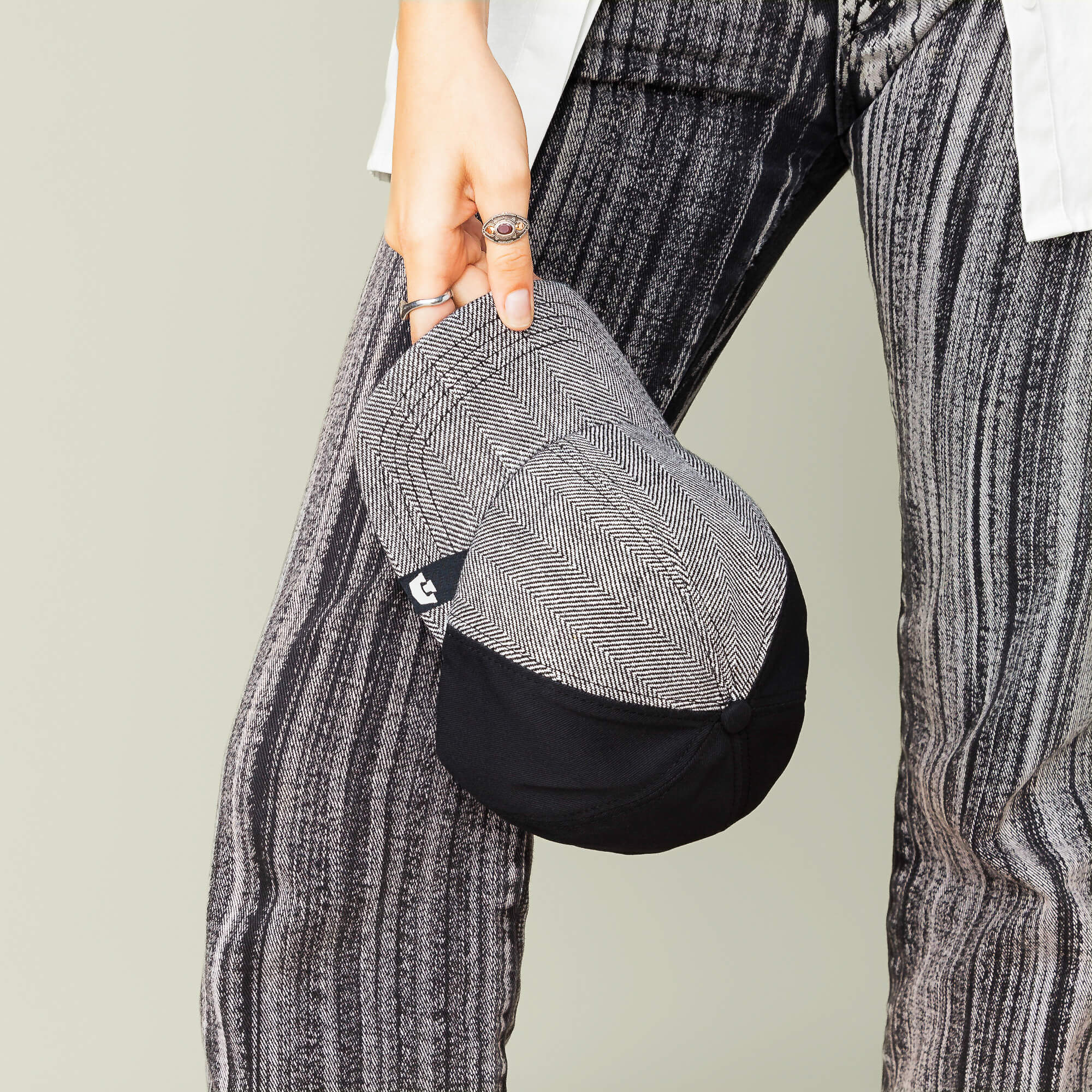 A person holds the Goorin Bros. Zig Zag Tailored Baseball hat, featuring a black and gray herringbone pattern, next to their striped pants; only the lower body and hand are visible.