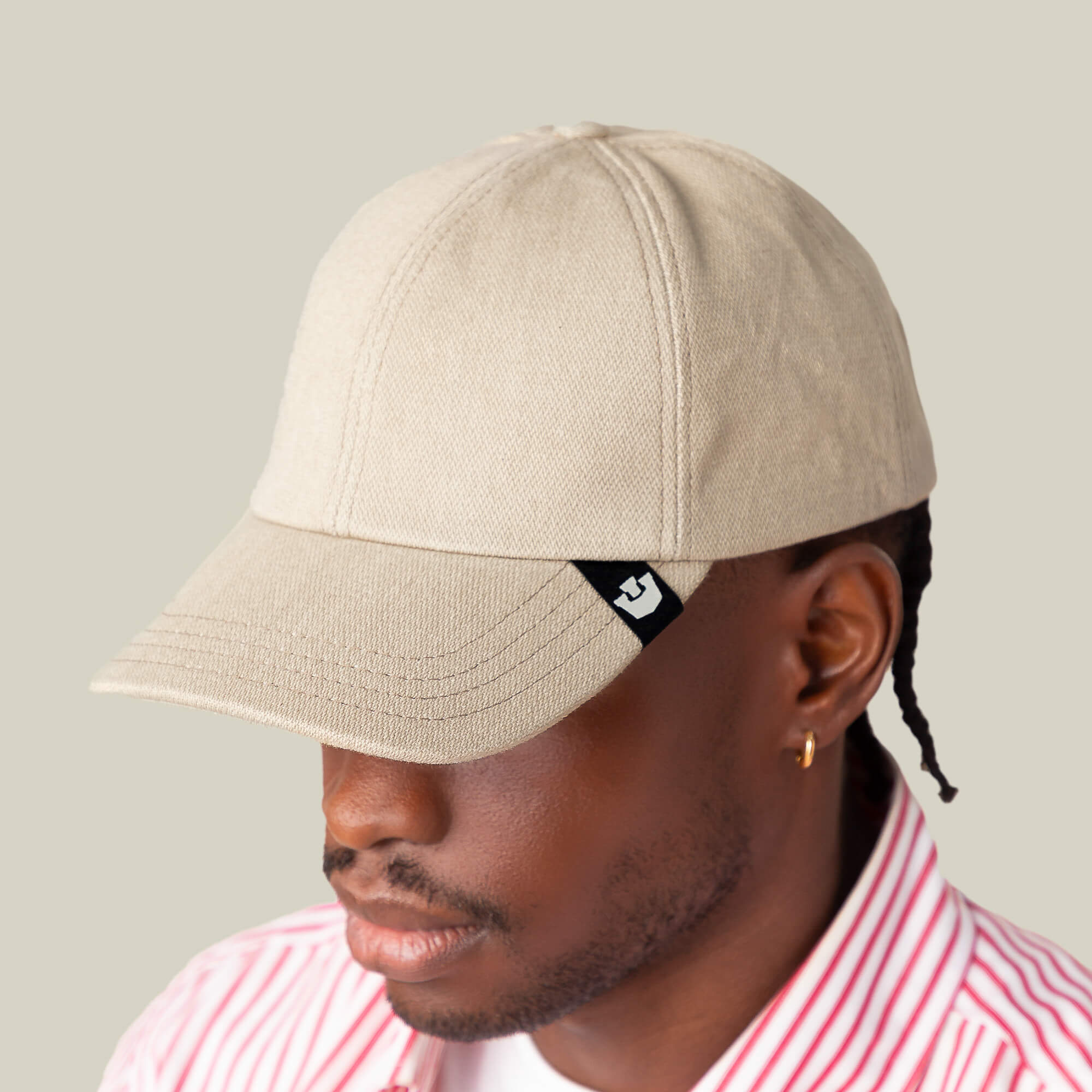 A person exudes subtle confidence wearing the Goorin Bros. Noisy Tailored Baseball cap in beige, featuring a small black logo tag, paired with a pink-and-white striped shirt against a plain light background.