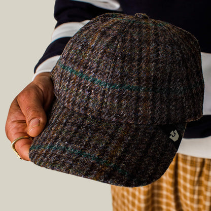 A person holds the Goorin Bros. Cozy Tailored Baseball cap in a muted plaid colorway with a small black tag, wearing a striped sleeve shirt and patterned pants.