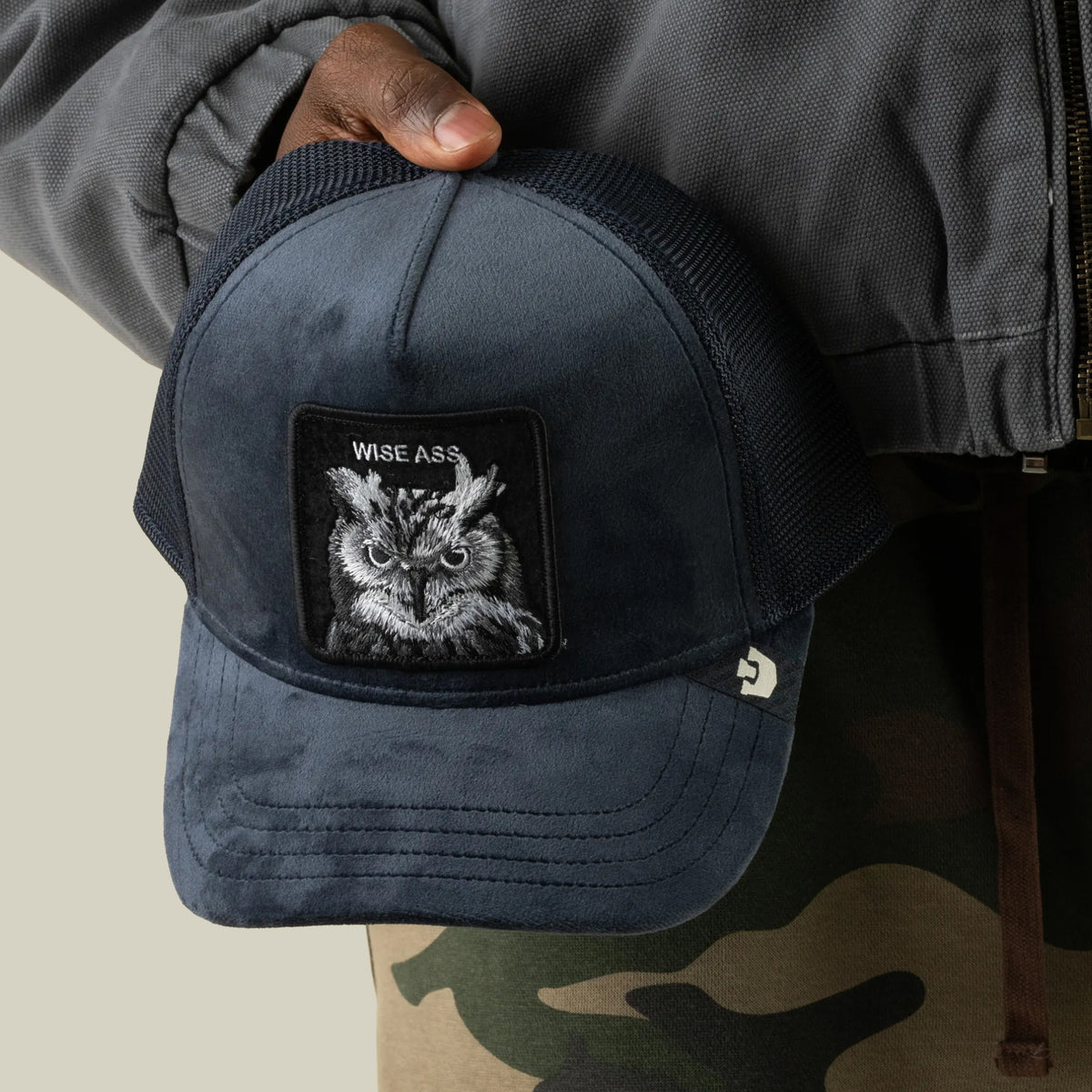 A person in a gray jacket and camo pants holds the Goorin Bros. Velour Wise Ass trucker hat, a navy blue mesh-back cap featuring an owl patch and the text WISE ASS on the front.
