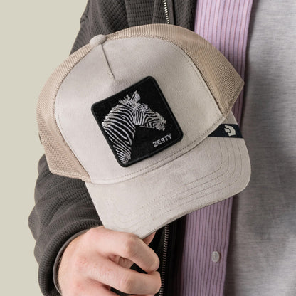 A person holds the Goorin Bros. Velour Zesty trucker hat in beige and tan, featuring a black patch with a white zebra head and the word “ZESTY,” its design accentuated by subtle monochrome stripes.