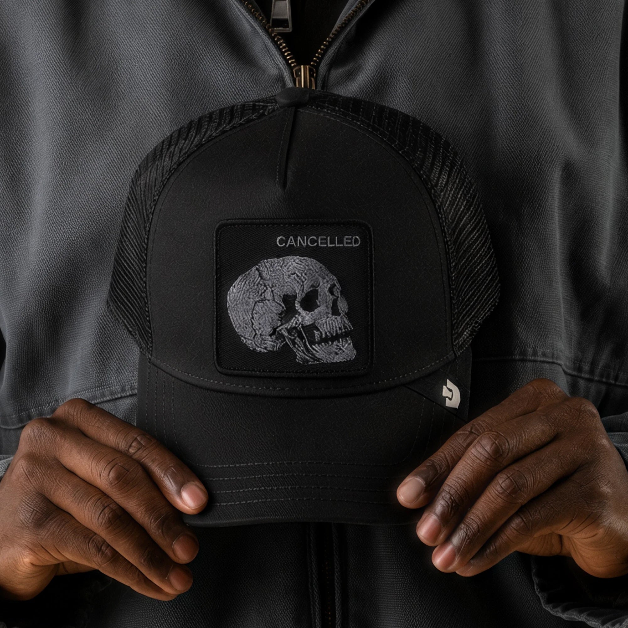 A person holds the Goorin Bros. Tonal Skull collectible matte trucker hat, which features a patch displaying a cracked skull with the word “CANCELLED” above it.