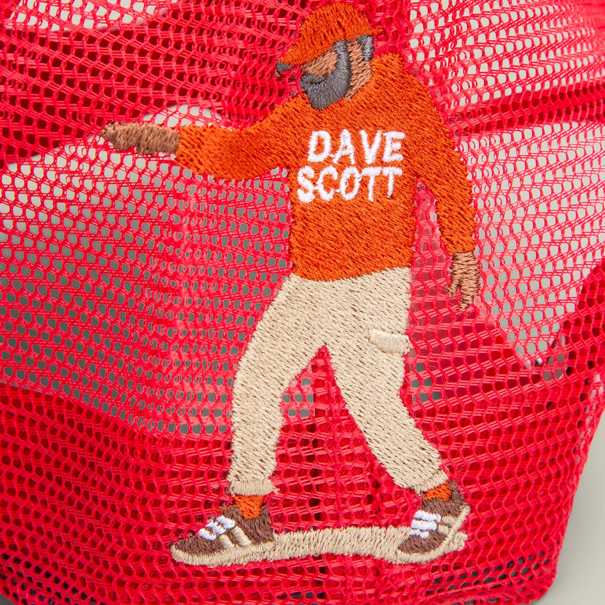The Dave Scott Boom Goat by Goorin Bros. features an embroidered hip hop legend in a red BOOM hat, orange sweater, beige pants, and sneakers on red mesh fabric.