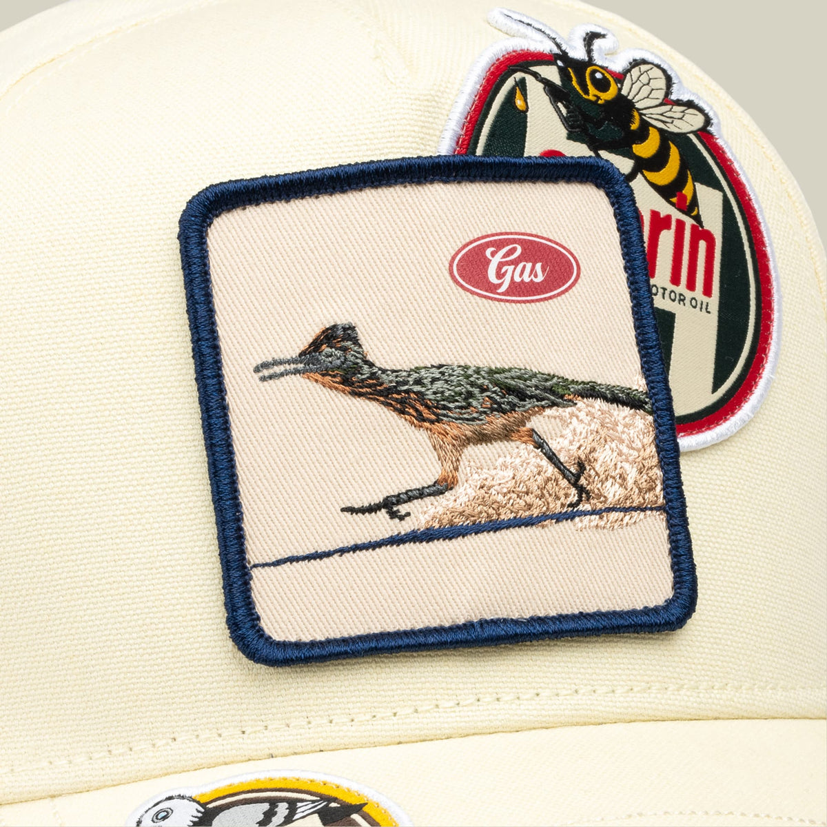 The Goorin Bros. Crude Guzzler trucker hat features a cream-colored front panel with navy trim, embroidered with a roadrunner and Gas logo, paired with embroidered bee and oil can patches on the back in red, yellow, and black.
