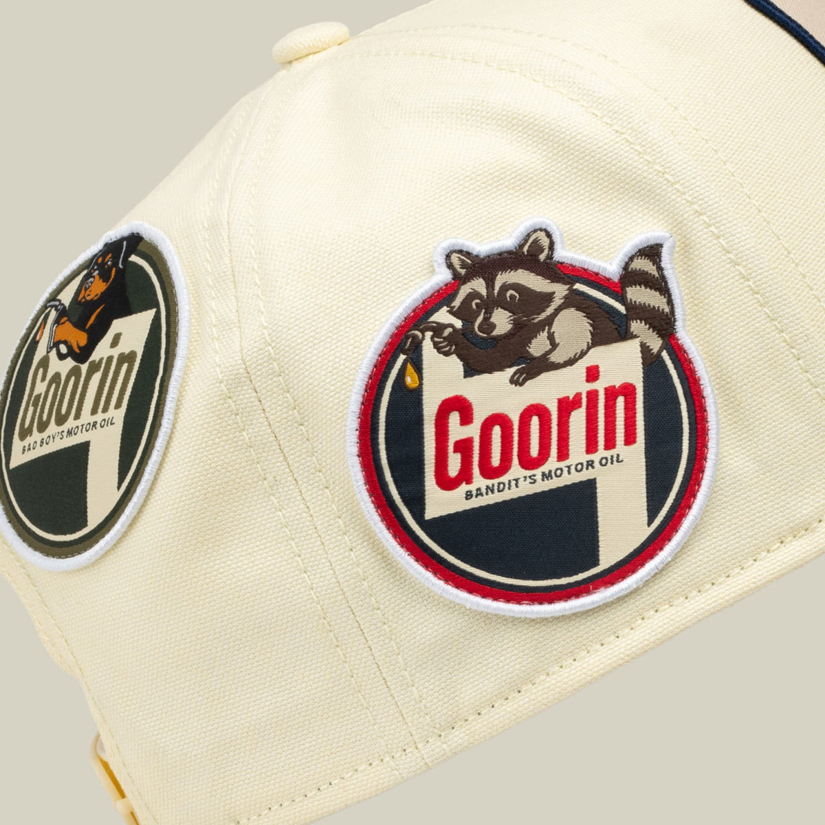 The Goorin Bros. Crude Guzzler is a cream-colored trucker cap featuring embroidered vintage patches, including a raccoon mascot and Goorin Bandit's Motor Oil branding in red, navy, and gold tones.
