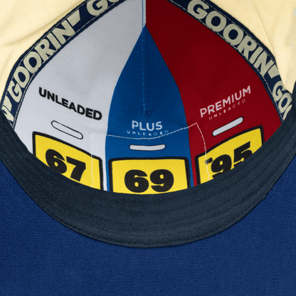 The Crude Guzzler by Goorin Bros is a navy blue snapback cap featuring a fuel gauge graphic design with white, red, and yellow sections displaying octane ratings, displayed flat lay against a blue surface.