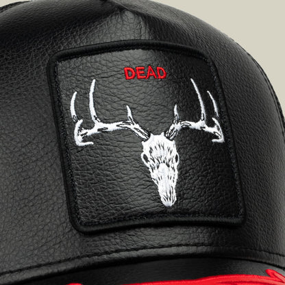 The Dead Bones Trucker by Goorin Bros. is a black leather trucker hat featuring an embroidered white deer skull with antlers and red "DEAD" text on a patch, displayed in a close-up studio shot.